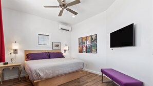 Ein Schlafzimmer mit Bett, Deckenventilator, Klimaanlage, Fernseher, einer Sitzbank und Kunstwerken an der Wand.