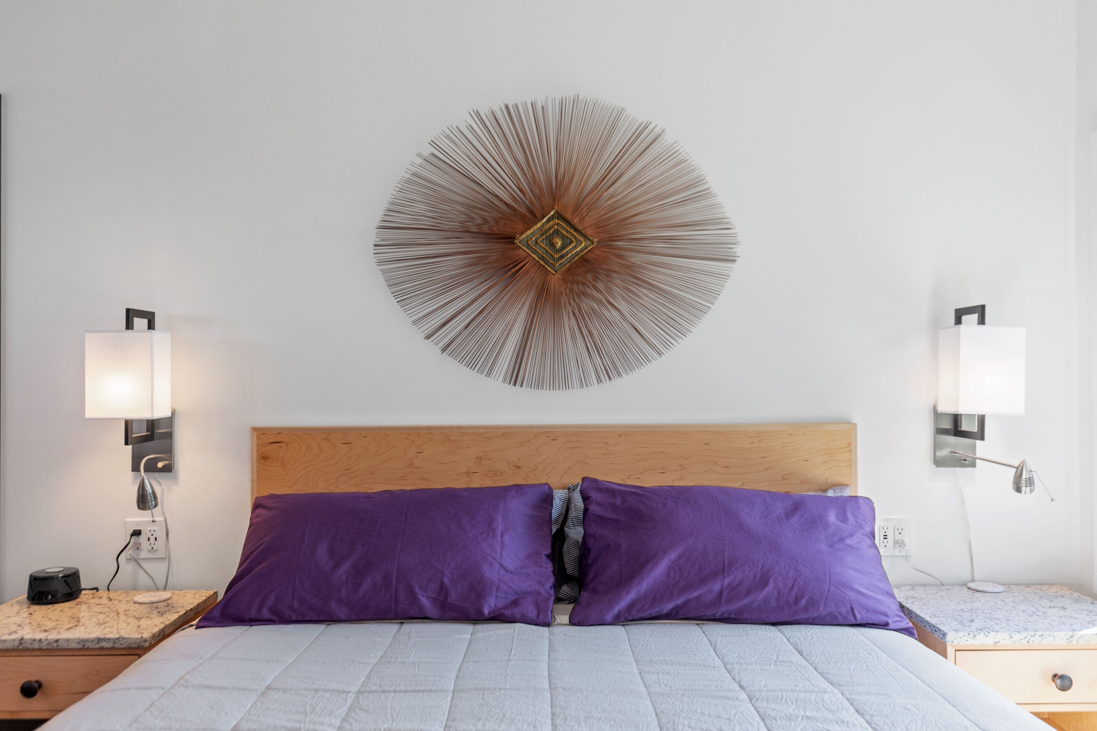 A neatly arranged bedroom with a wooden headboard, two purple pillows, a round wall decoration, and bedside lamps.