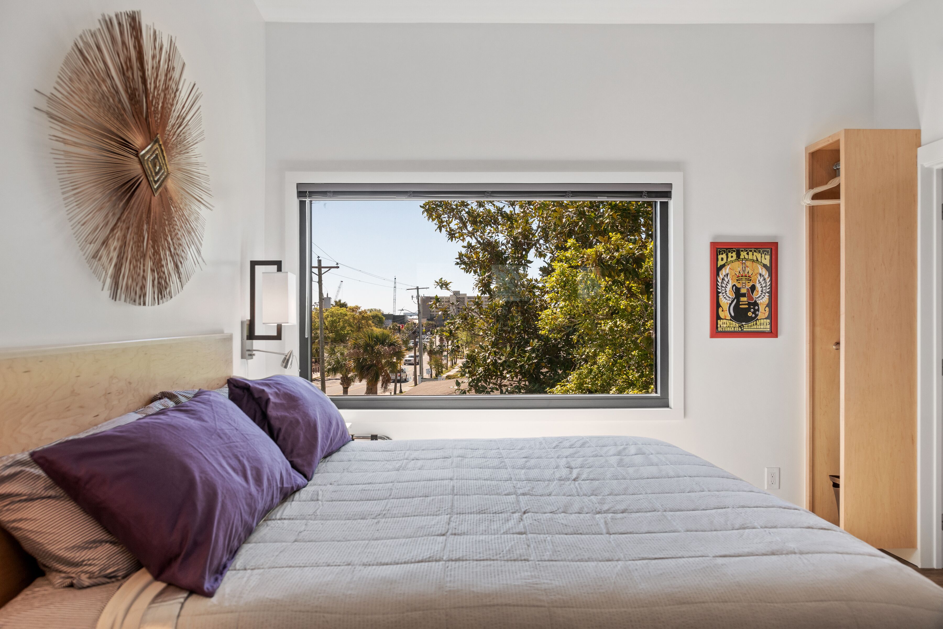 Une chambre avec une grande fenêtre, un lit recouvert d’une couette blanche, un oreiller violet, une œuvre d’art murale et une armoire en bois.