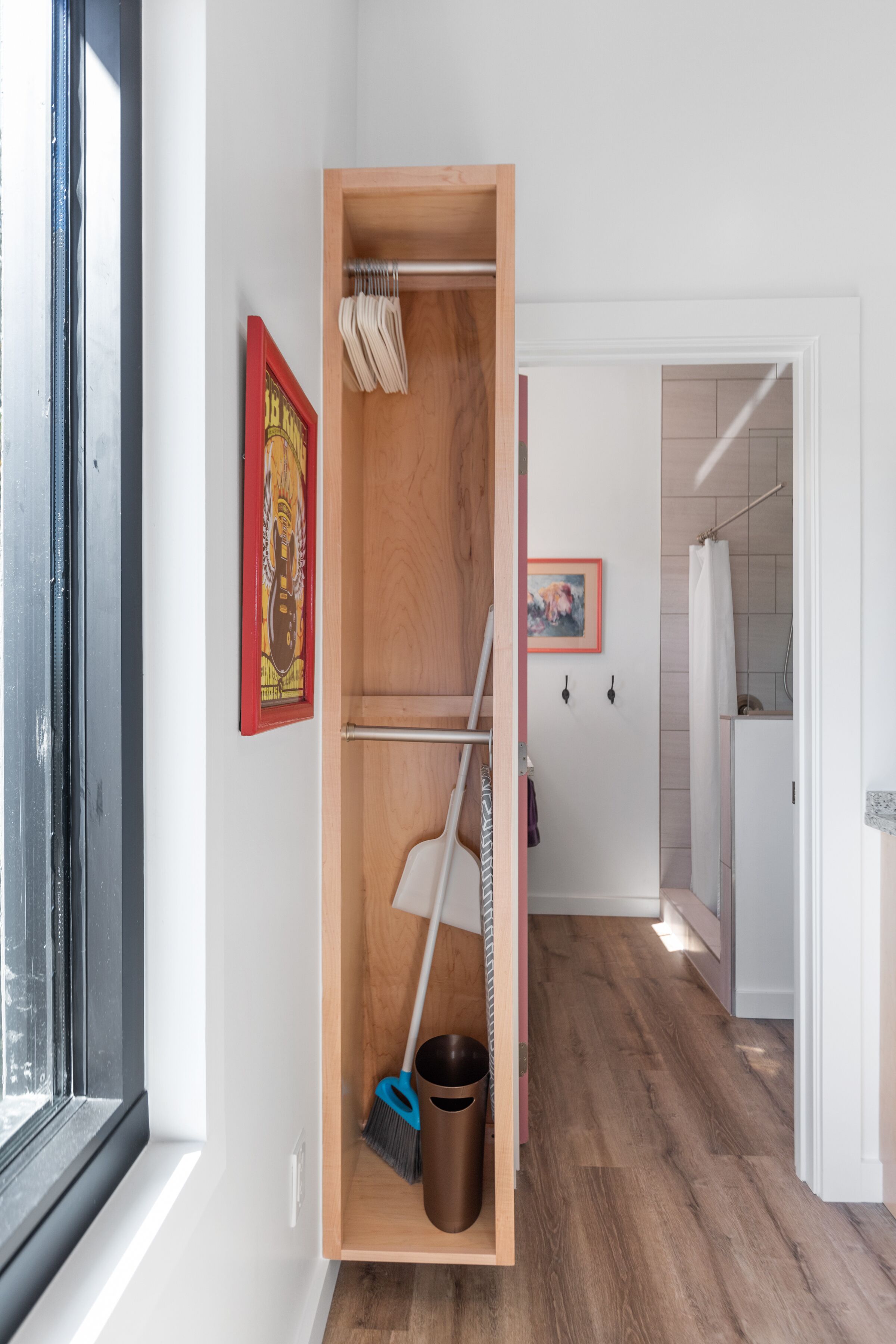 Un couloir étroit avec une étagère en bois contenant un balai, une pelle à poussière et une poubelle. On aperçoit une fenêtre et une porte donnant sur une salle de bain.