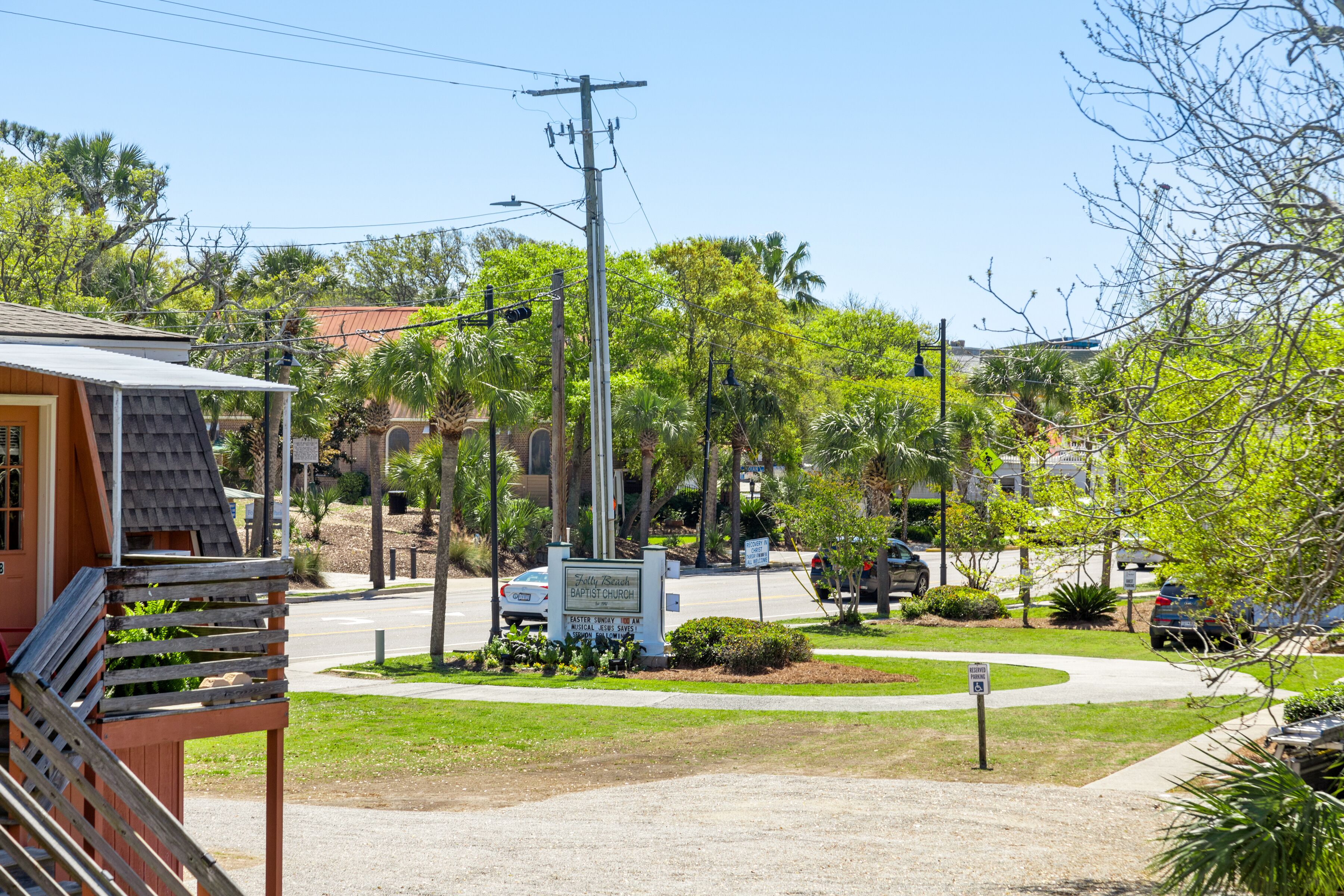 A church with a signboard, surrounded by greenery and a parking area.