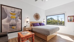 A bedroom with a bed, a nightstand, a table, a window, and a wall art piece.