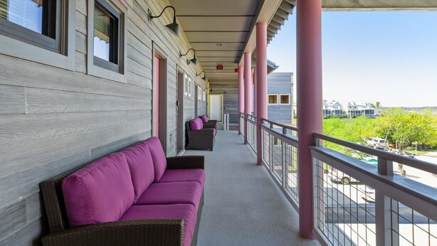 Een overdekt balkon met een rij rieten banken en een hekwerk, met uitzicht op een straat met gebouwen en begroeiing.