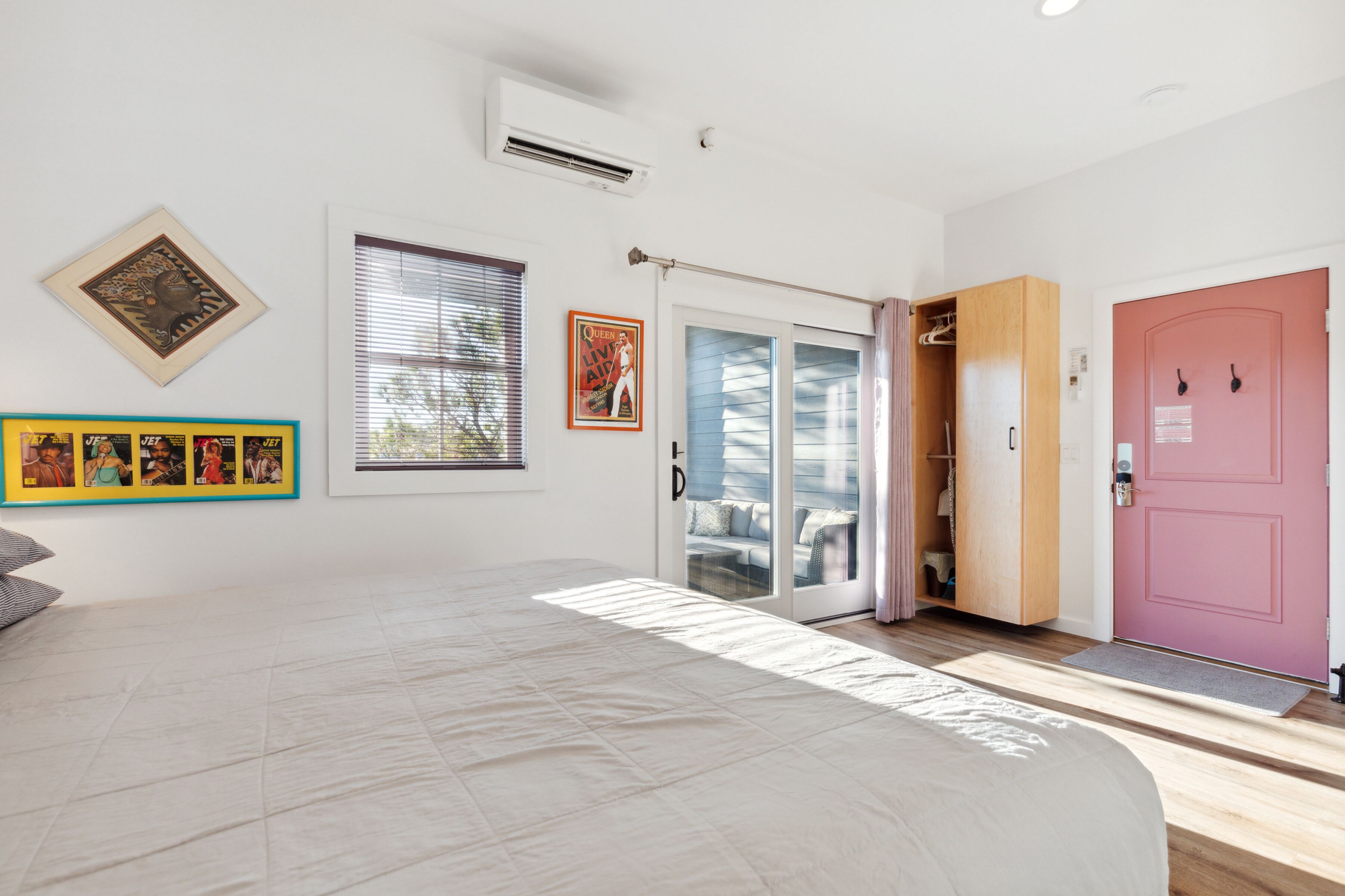 Une chambre avec un lit, une porte-fenêtre coulissante donnant sur un balcon, une porte rose et un climatiseur fixé au mur.