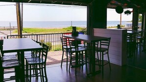 Un área de comedor al aire libre con mesas y sillas, vistas al mar y una sección cubierta con mostrador y sillas.