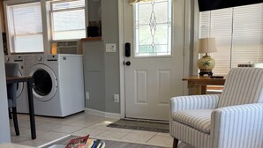 Un salón con un sillón a rayas, una mesa de centro con libros, una lavadora y un televisor al fondo.