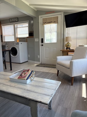 A living room with a striped armchair, a coffee table with books, a washing machine, and a TV in the background.