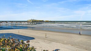 A beach with a pier extending into the ocean, a person walking on the sand, and a clear sky.