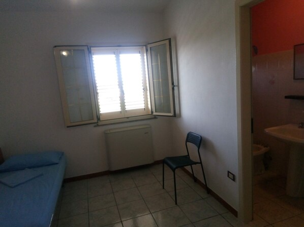 A small room with a bed, a chair, a window with blinds, and a bathroom with a sink. - Double Room for Rent with Private Bathroom (Marina di Montenero di Bisaccia)
