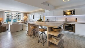 A modern kitchen with a central island, bar stools, a microwave, oven, and a large window overlooking a cityscape. - Luxurious 1/1 Ocean View Condo 1416 - Located at 1 Hotel & Homes South Beach (Miami Beach)
