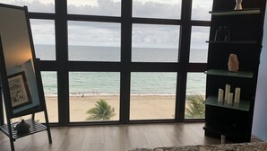 Una habitación con grandes ventanales que dan a una playa, una cama, un estante con objetos decorativos y un cuadro en un atril.
