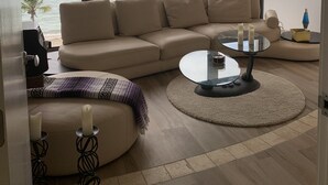 Un salón con un ventanal grande con vistas al mar, un sofá color beige, una mesa de centro de cristal y una alfombra estampada.