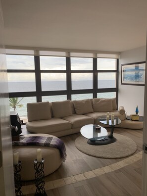 A living room with a large window overlooking the sea, a beige sofa, a glass coffee table, and a patterned rug.