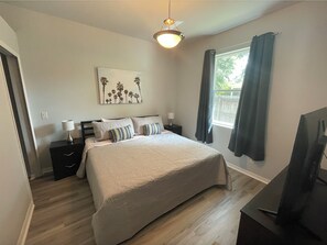 Une chambre à coucher avec un lit, des tables de chevet, une lampe, un tableau, une fenêtre avec des rideaux et un téléviseur.