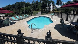 Piscina com cadeiras de praia e guarda-sóis, cercada por uma grade e árvores.