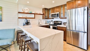 A modern kitchen with a marble island, stainless steel appliances, wooden cabinets, and bar stools. - Exceptionally Remodeled Oceanfront, Sunsets,  A/C, King bed Hale Kai O Kihei (Kihei)
