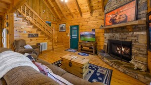 A log cabin interior with a stone fireplace, wooden staircase, and cozy seating area. - Gypsy Road | Secluded Wears Valley Cabin with Hot Tub, Arcade & Pool Access (Sevierville)