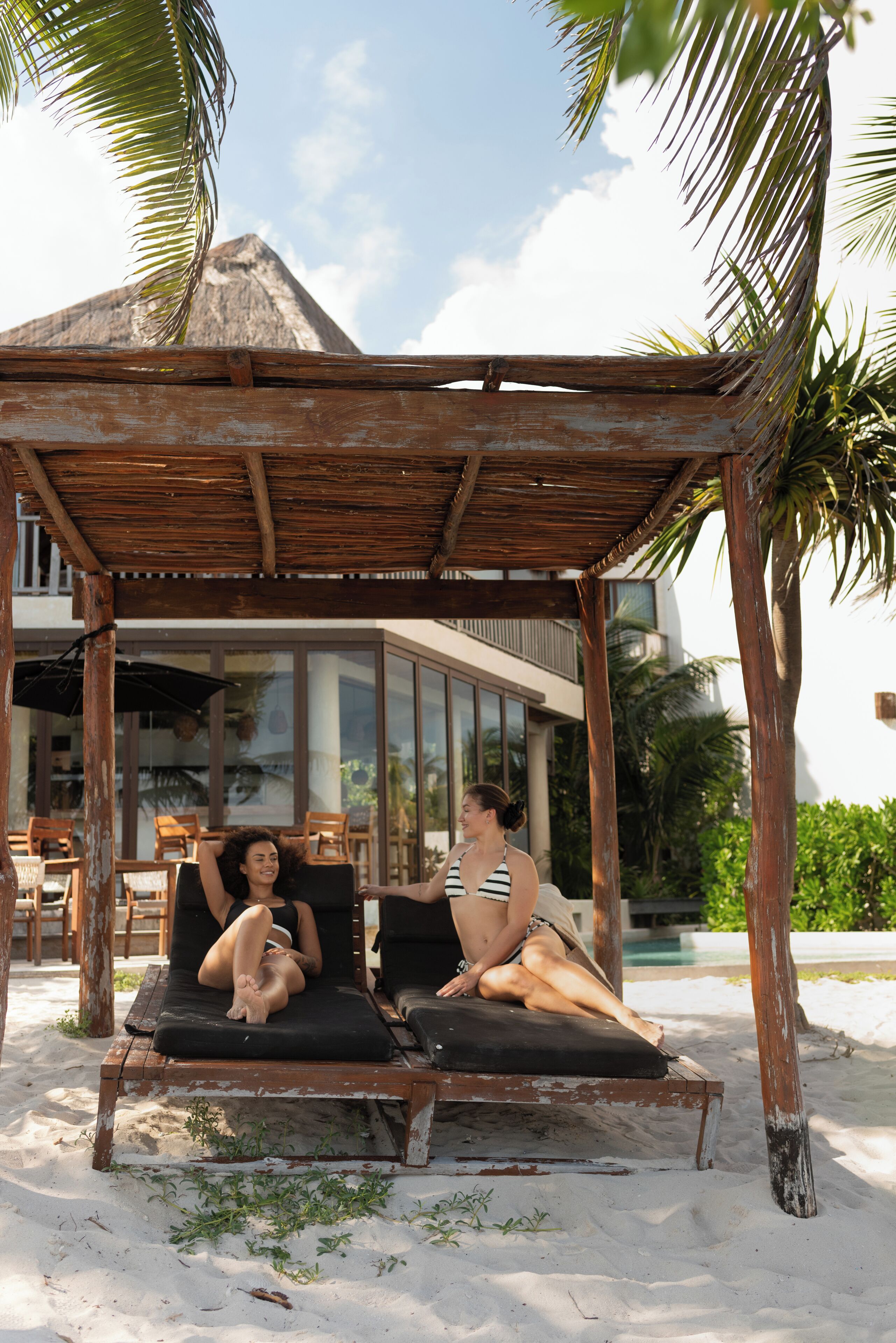 Zwei Personen entspannen in Holzliegen unter einer Holzpergola an einem Sandstrand, mit einem Gebäude und Palmen im Hintergrund.