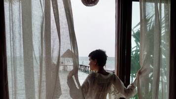 Una persona de pie junto a una ventana con cortinas traslúcidas, mirando hacia una playa con una estructura de techo de paja.
