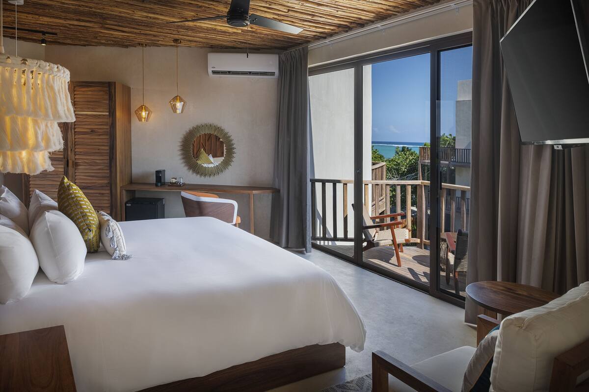A bedroom with a large bed, a desk, and a balcony with a view of the ocean.