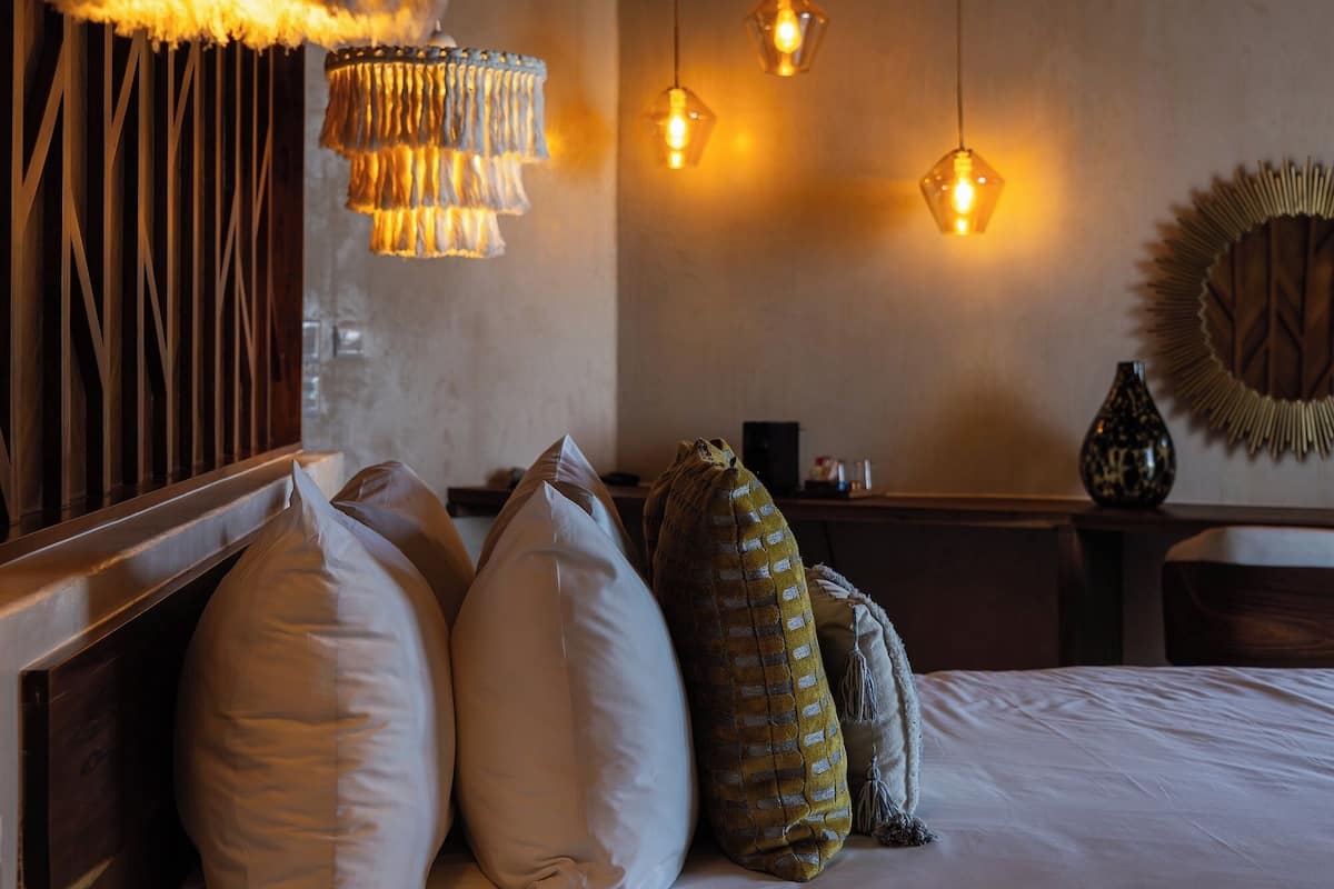A hotel room with a bed, pillows, a remote control, a wall-mounted mirror, and hanging pendant lights.