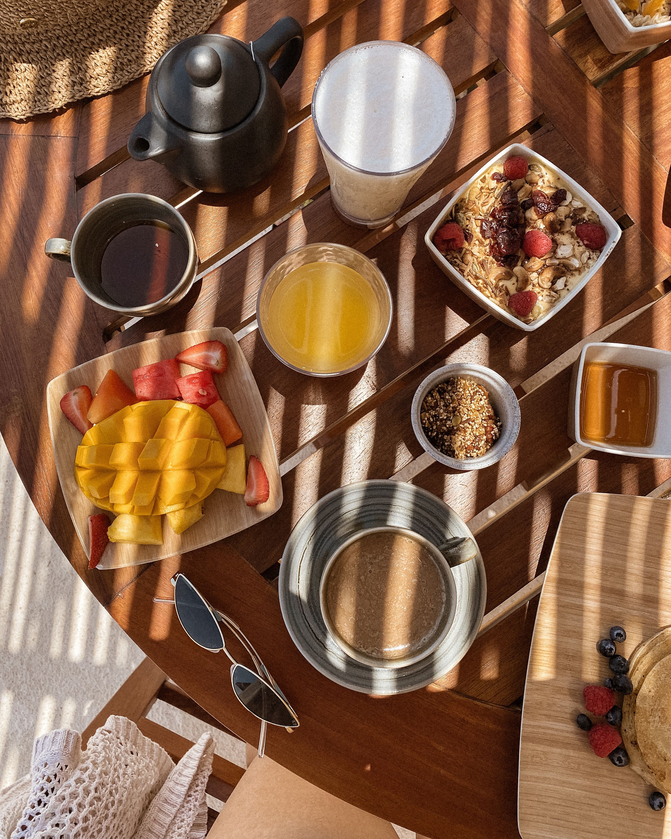 A breakfast table with a teapot, cups, a bowl of fruit, a plate of cereal, and a cup of coffee.