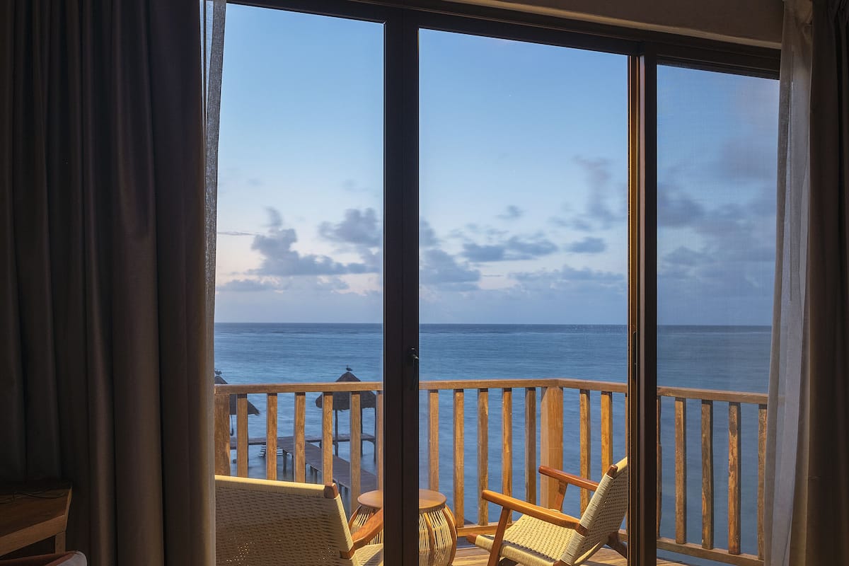 A room with a large sliding glass door leading to a balcony with a view of the ocean. There is a bed, a chair, and a small table on the balcony.