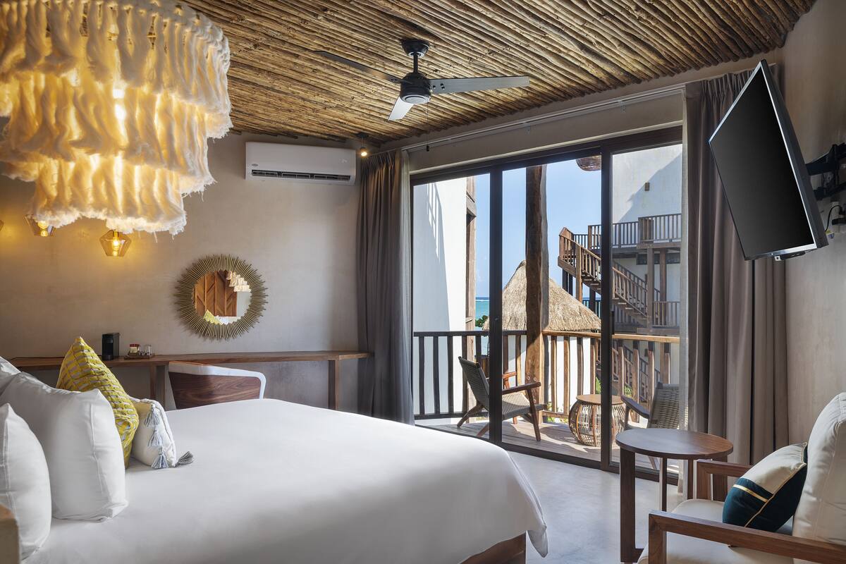 A hotel room with a large bed, a ceiling fan, a TV mounted on the wall, and a balcony with a view of the sea.