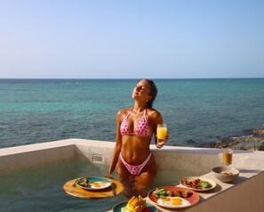 A woman in a bikini at a poolside table with breakfast items and a glass of orange juice, overlooking a clear blue sea.