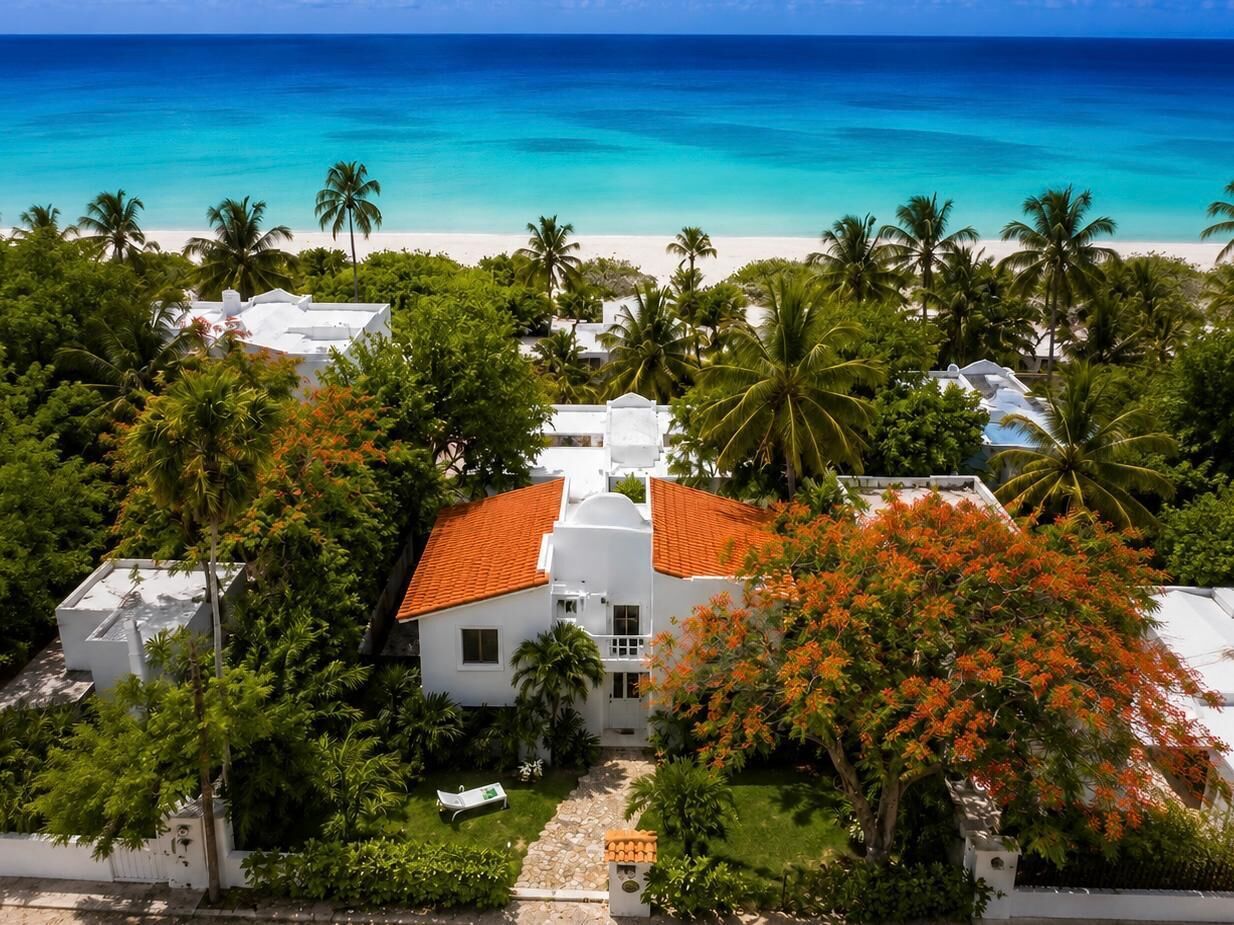 A white building with a red roof surrounded by lush greenery and palm trees, overlooking a beach.