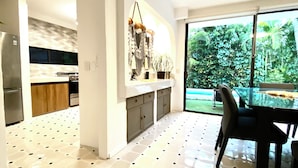 Una cocina moderna con una isla grande, un comedor con mesa de vidrio y vistas a un jardín exuberante a través de amplios ventanales.