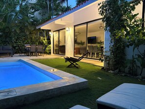 Ein moderner Poolbereich im Freien mit Liegestuhl, Essplatz und einer verglasten Terrasse.