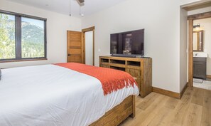 A bedroom with a large bed, a TV on a wooden stand, a wooden door, and a view of the outdoors.