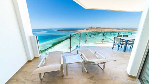A balcony with two lounge chairs and a table, overlooking a beach and turquoise waters.