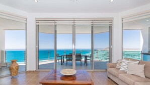 A room with a large sliding glass door leading to a balcony with a view of the ocean. There is a wooden table with a bowl on it, a beige sofa, and a vase on the floor.