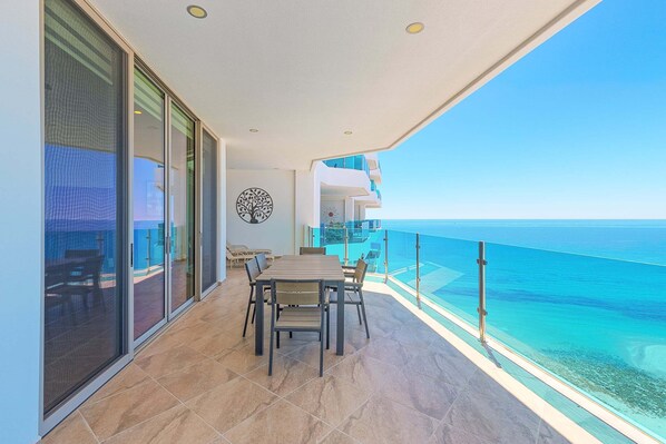 A balcony with a view of the ocean, a dining table with chairs, and a glass door leading to an outdoor area.