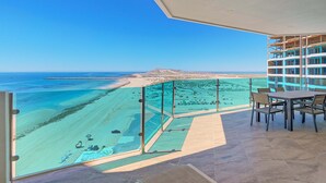 A balcony with a view of a beach and ocean, a dining table with chairs, and a bookshelf.