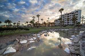 Un complexe hôtelier doté d’une piscine entourée de rochers et de palmiers, d’un terrain de golf en arrière-plan et d’un immeuble à plusieurs étages avec des balcons.