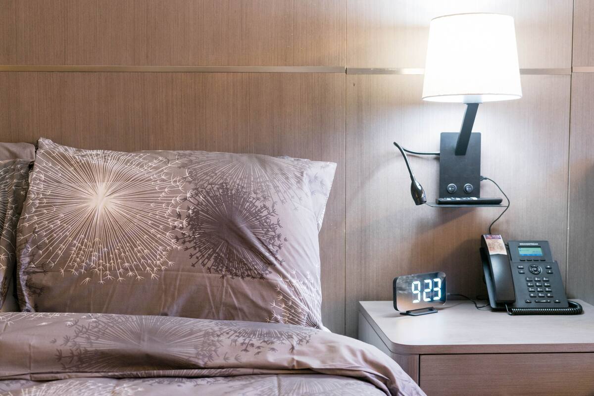 A hotel room with a bed, a nightstand with a lamp, a digital clock, and a telephone.