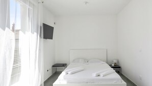 Camera d'albergo dal design minimalista con un ampio letto, una TV a schermo piatto montata a parete, un comodino con lampada e tende leggere alla finestra.