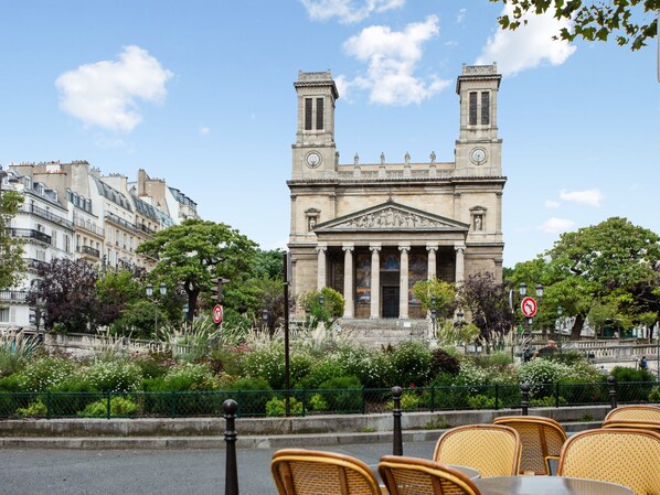 Un bâtiment historique à l’architecture classique, orné de colonnes et de statues, entouré de verdure et de bâtiments urbains.