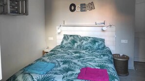 Makuuhuoneessa on sängyssä trooppisten lehtien kuvioitu päällinen, seinälle kiinnitetty hylly, jossa on teksti ”OCEAN”, pieni yöpöytä ja roskakori.