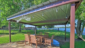 Un área de picnic techada con una mesa y sillas, un cartel que dice "Welcome to the Lake" y una vista de un lago y árboles al fondo.