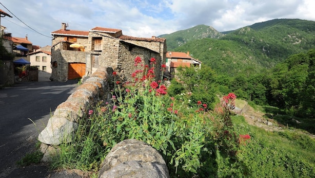 Un bâtiment en pierre surmonté d’un toit de tuiles rouges, entouré d’une végétation luxuriante et avec des montagnes en arrière-plan.