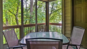 A covered outdoor area with a table and chairs, surrounded by wooden railings and large windows offering a view of trees. - Watefront Branson Condo w/ Screened-In Balcony! (Branson)