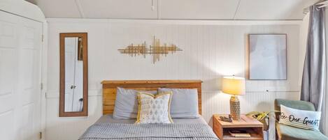 A bedroom with a wooden headboard, a bed with grey bedding, a bedside table with a lamp, a chair, and a mirror on the wall.