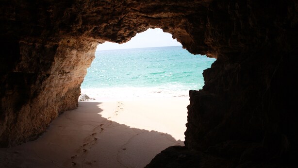 Vista de una playa a través de la entrada de una cueva, con un sendero arenoso que lleva al agua.