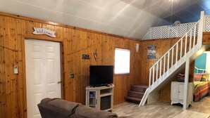 A cozy living room with a brown couch, a TV on a small stand, a staircase with a white railing, and a 'Welcome' sign on the door.