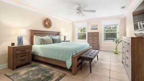 A bedroom with a bed, bedside tables, a bench, a ceiling fan, and a view of the outdoors through windows. - Complete Searenity by Distinctive Beach Rentals (Palm Coast)