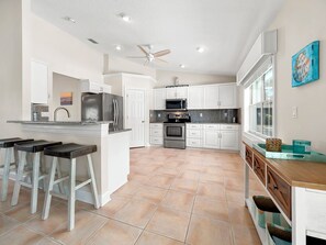 A modern kitchen with white cabinets, a stainless steel refrigerator, a microwave, and a stove. There are bar stools and a ceiling fan. - Complete Searenity by Distinctive Beach Rentals (Palm Coast)